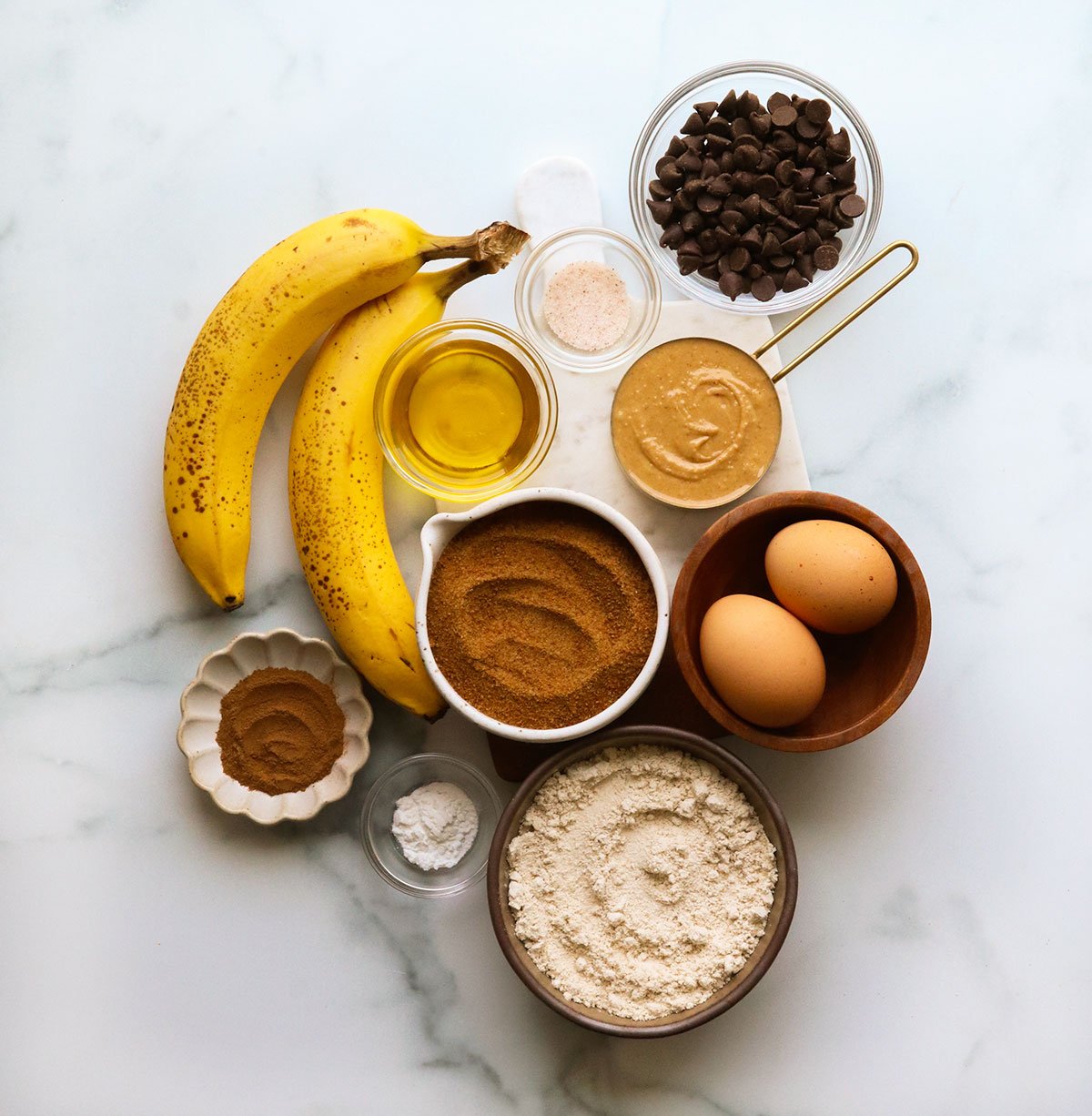 bananas, coconut sugar, oat flour, eggs, peanut butter, and cinnamon on a white surface.