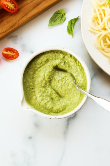 zucchini pesto in a white bowl next to a plate of pasta and cherry tomatoes.